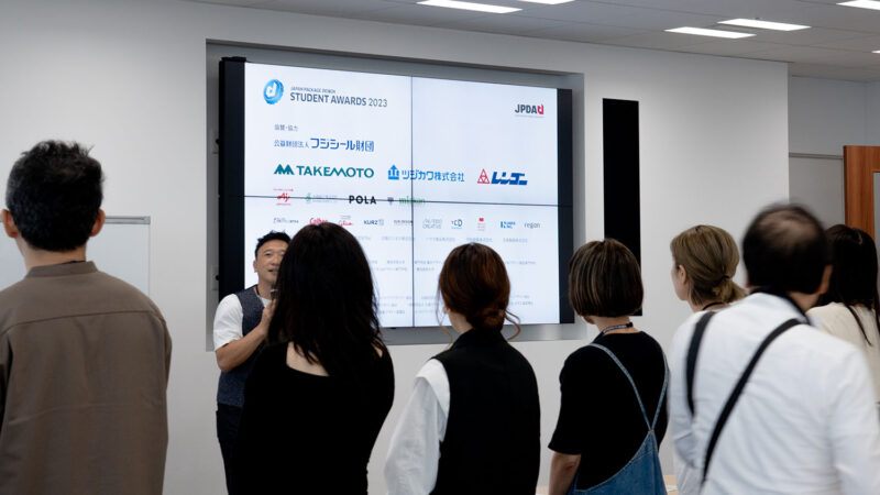 男性が前に立ち、まわりに人がいる。男性の後ろのスクリーンにスポンサー名が表示されている。